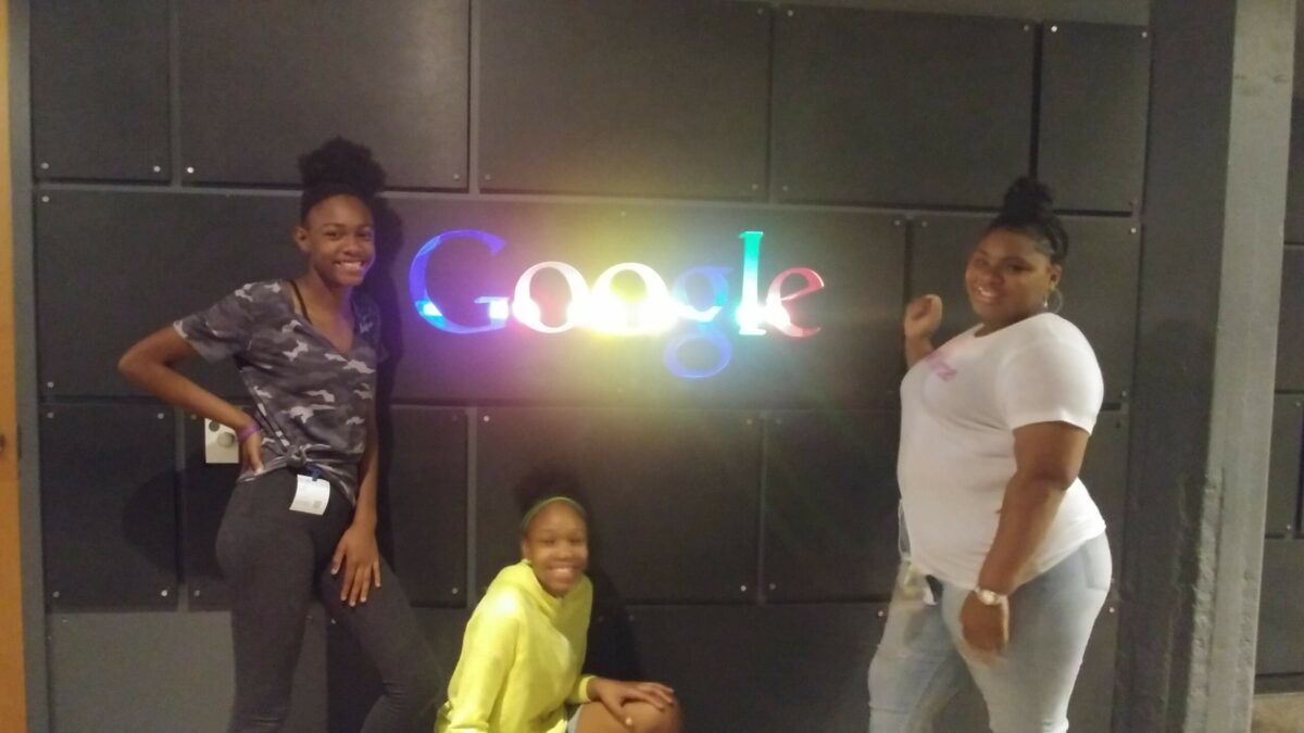 Three youth posing next to and underneath the text "Google" with each letter lit up in blue, red, yellow, blue, green, and red respectively