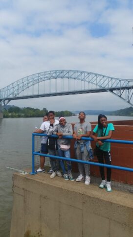Five youth posing behind a blue railing underneath a bridge overlooking a river