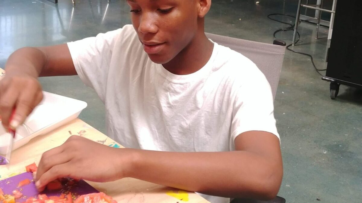 Youth dicing a tomato on a purple cutting board, with two youth having a discussion in the back corner in a classroom