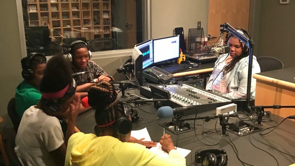 Four youth wearing headphones sitting behind microphones writing on pieces of paper, with another youth wearing headphones and sitting behind a mixing board in a radio studio