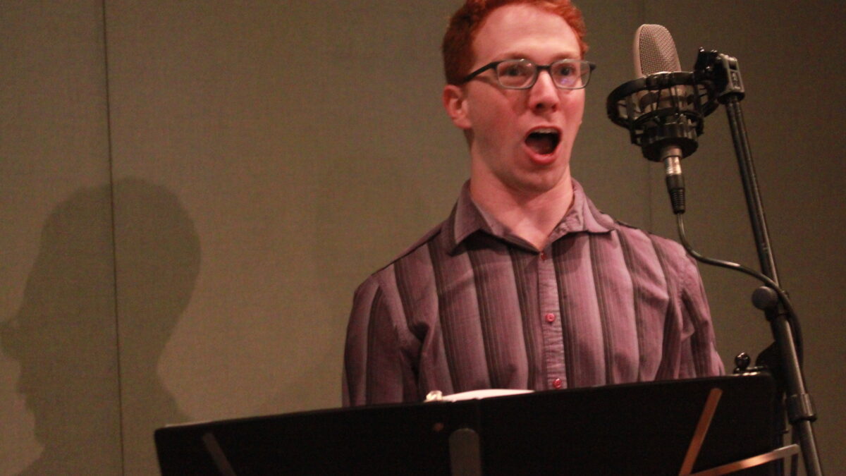 Adult singing into a microphone behind a music stand in a radio studio
