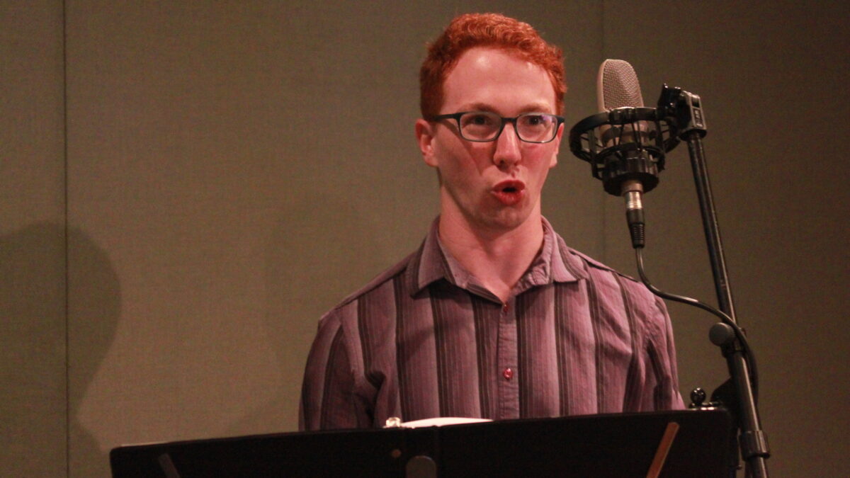 Adult singing into a microphone behind a music stand in a radio studio