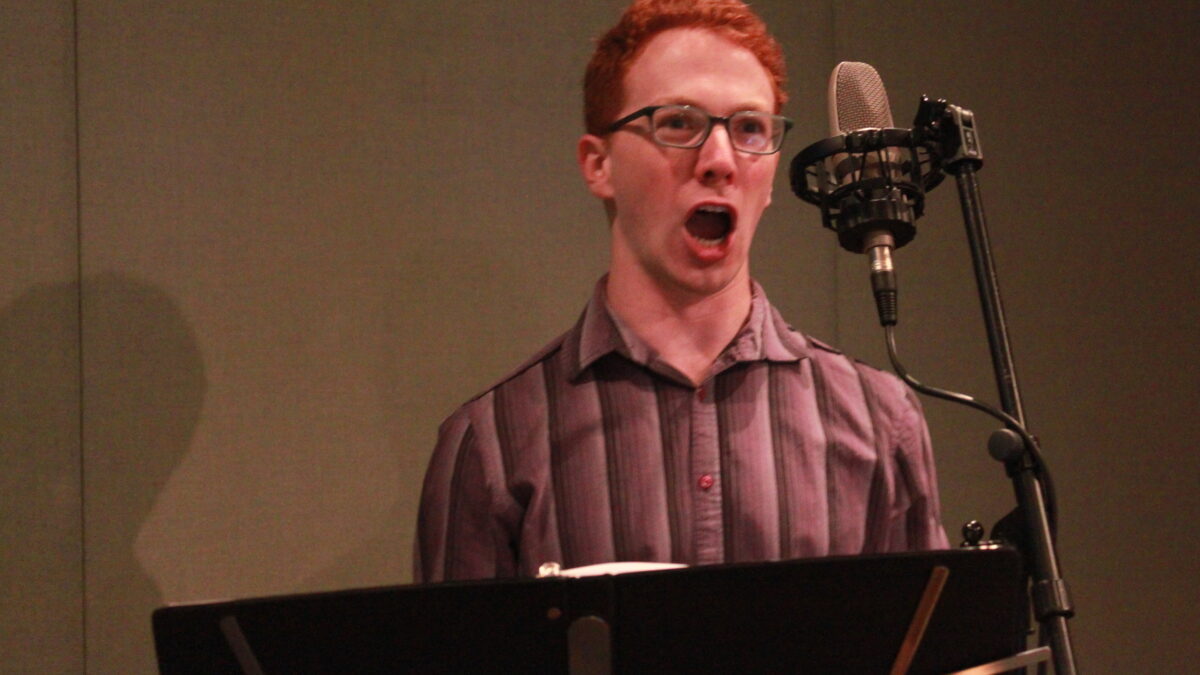 Adult singing into a microphone behind a music stand in a radio studio