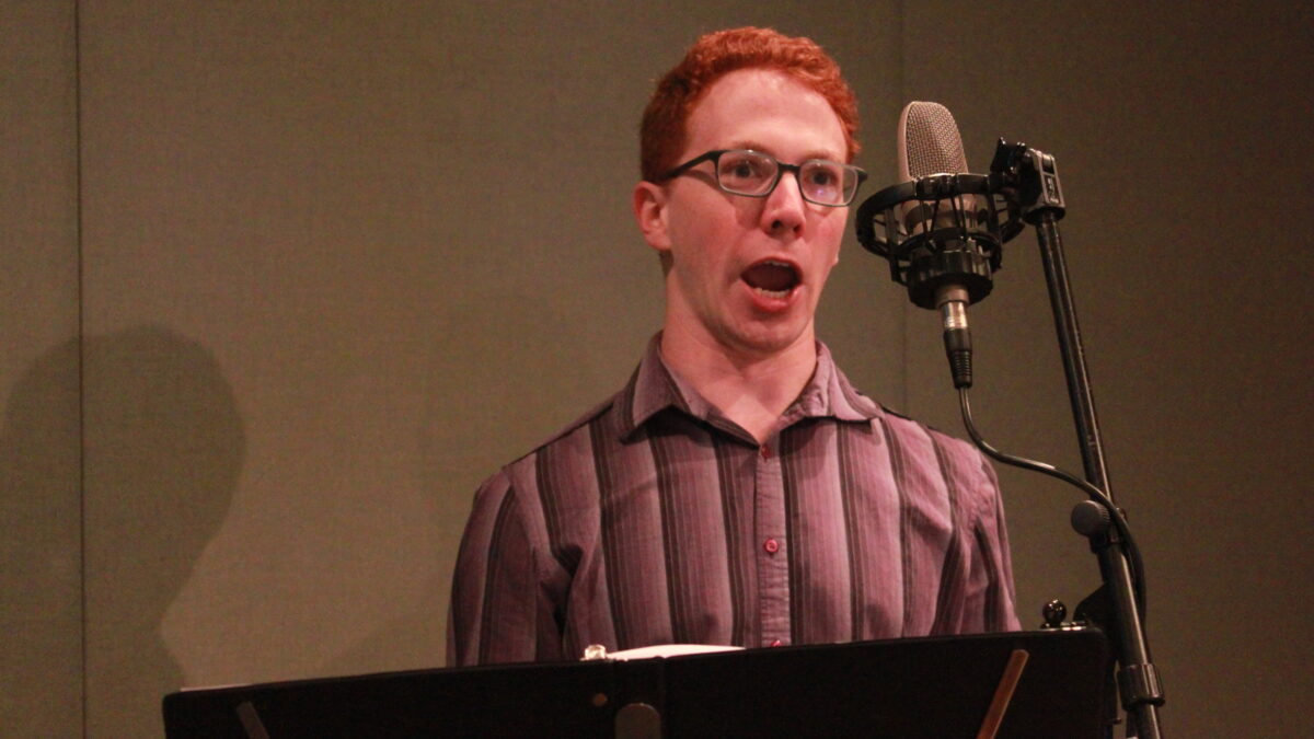 Adult singing into a microphone behind a music stand in a radio studio