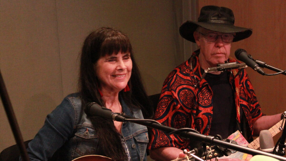 Adult playing mandolin and smiling and an adult playing cigar box guitar in a radio studio