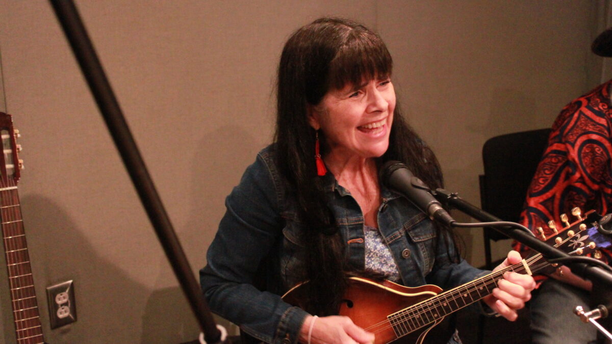An adult playing mandolin and singing into a microphone in a radio studio