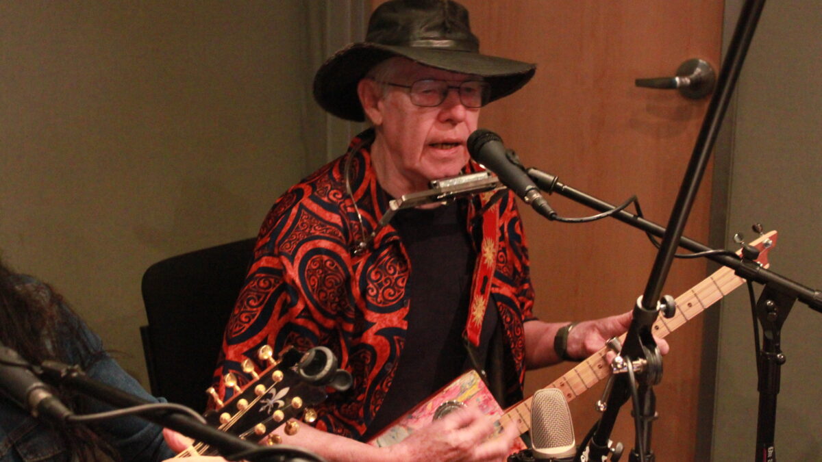 Adult singing into a microphone and playing a cigar box guitar in a radio studio