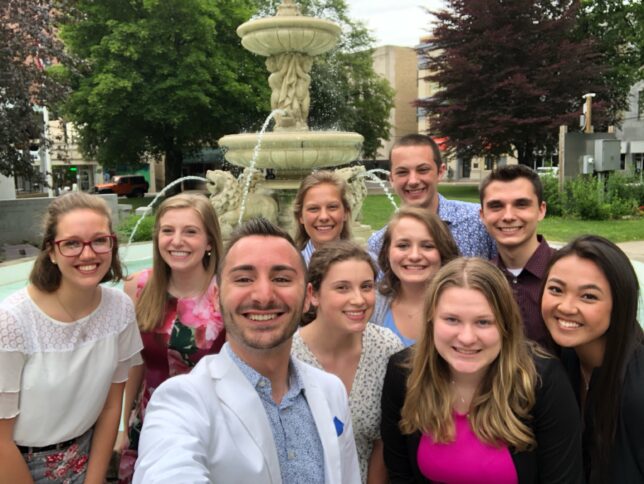 Ten young adults smiling, posing for a group photo in front of a green fountain.