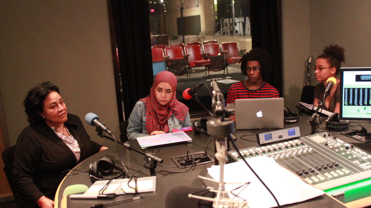 three youth looking an an adult, all sitting behind microphones and an open laptop and a closed laptop in a radio studio
