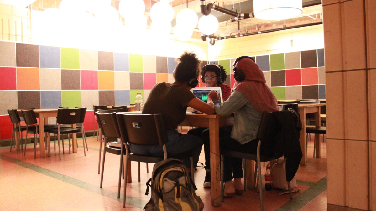 Three teens wearing headphones sitting around a table looking at a laptop displaying an audio waveform