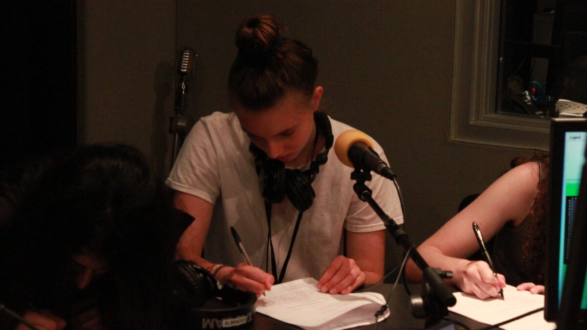 Two teens wearing headphones sitting behind a microphone writing on a piece of paper in a radio studio