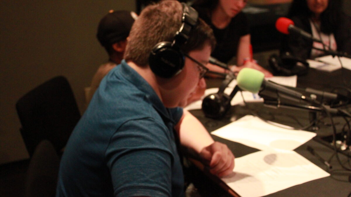 Four young adults sitting behind microphones and looking down at pieces of paper in a radio studio