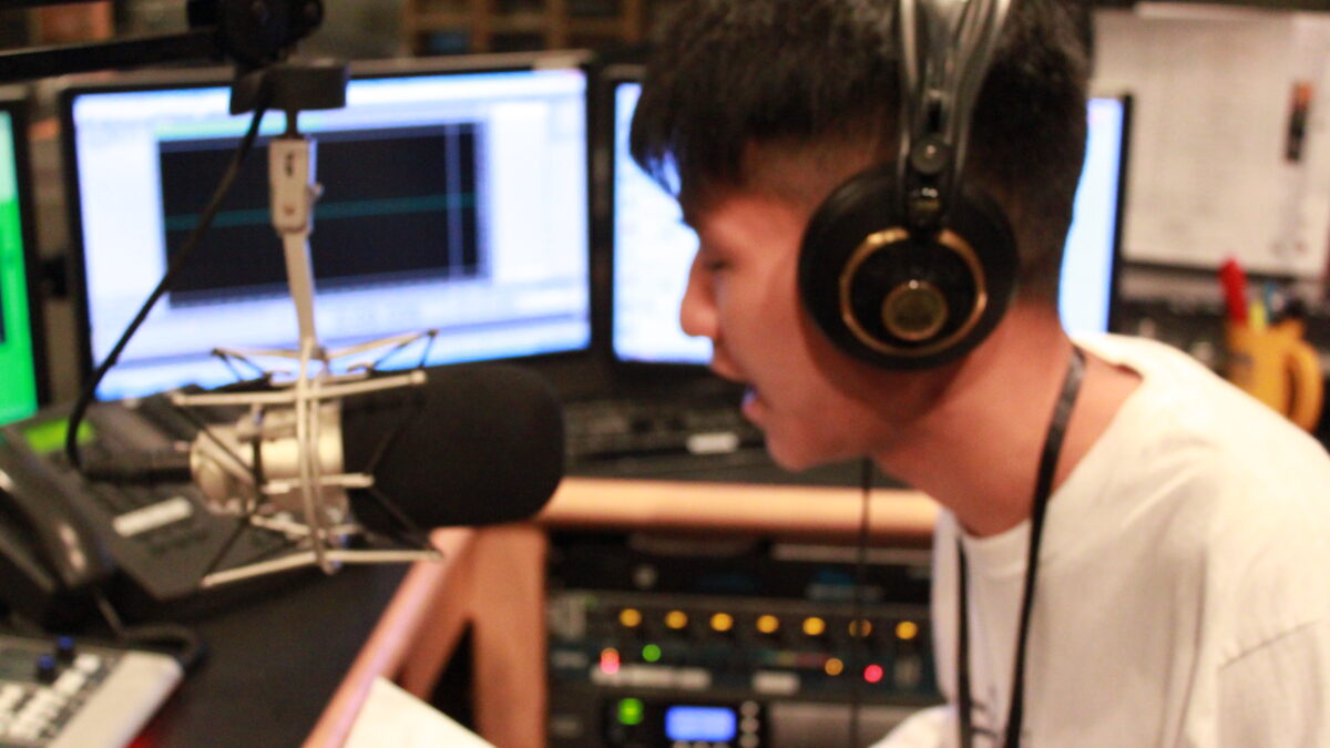 Teen wearing headphones speaking into a microphone sitting behind a mixing board and looking down at a piece of paper in a radio studio