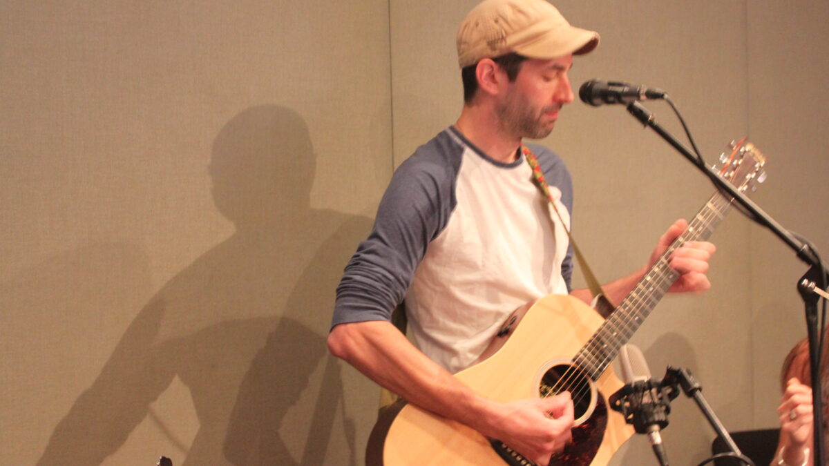 An adult playing guitar into a microphone in a radio studio