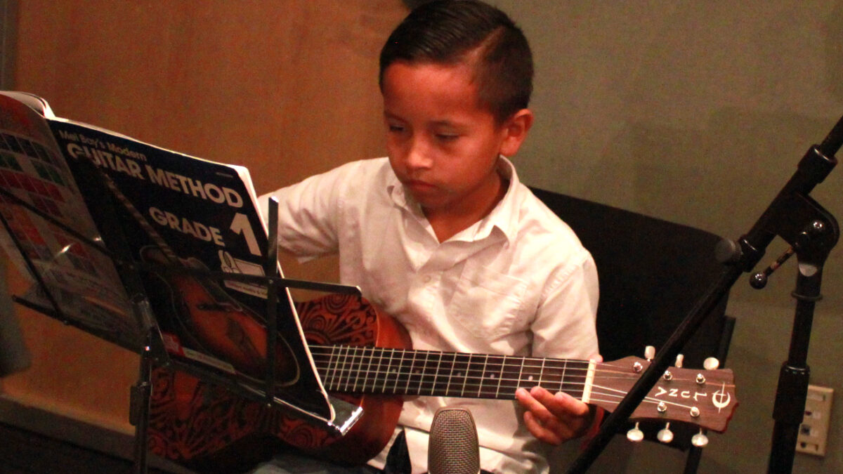 A child playing guitar while looking at sheet music on a music stand in a radio studio