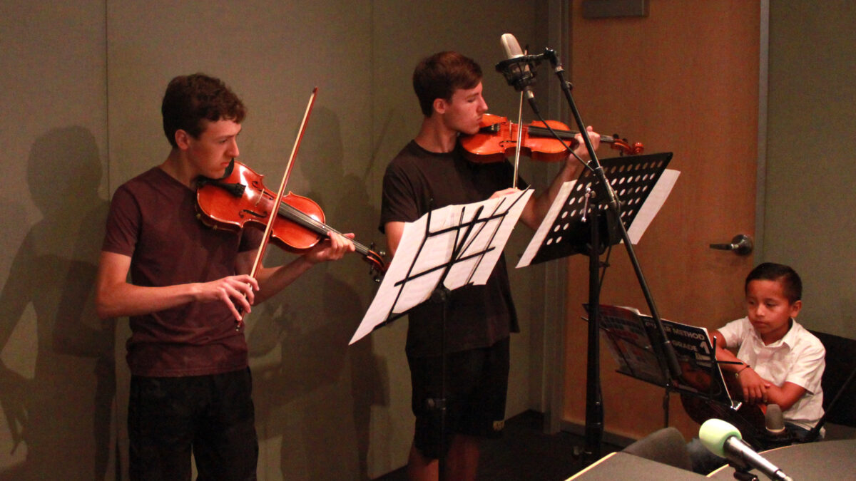 Three youth playing violin and looking at sheet music on music stands in a radio studio