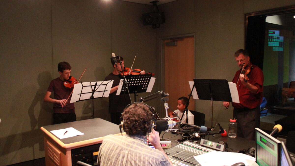 An adult playing violin and three youth playing violins, all looking at sheet music on music stands, while an adult wearing headphones behind a mixing board and a microphone listens in a radio studio