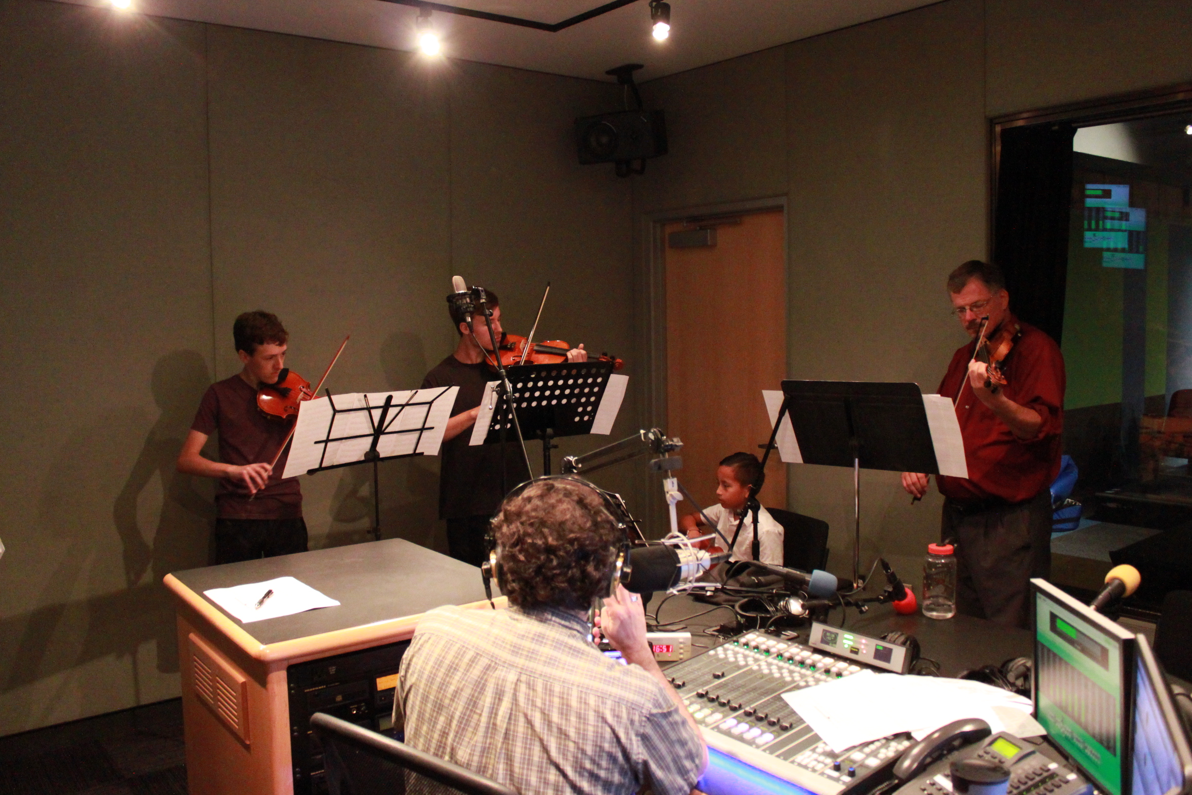 An adult playing violin and three youth playing violins, all looking at sheet music on music stands, while an adult wearing headphones behind a mixing board and a microphone listens in a radio studio