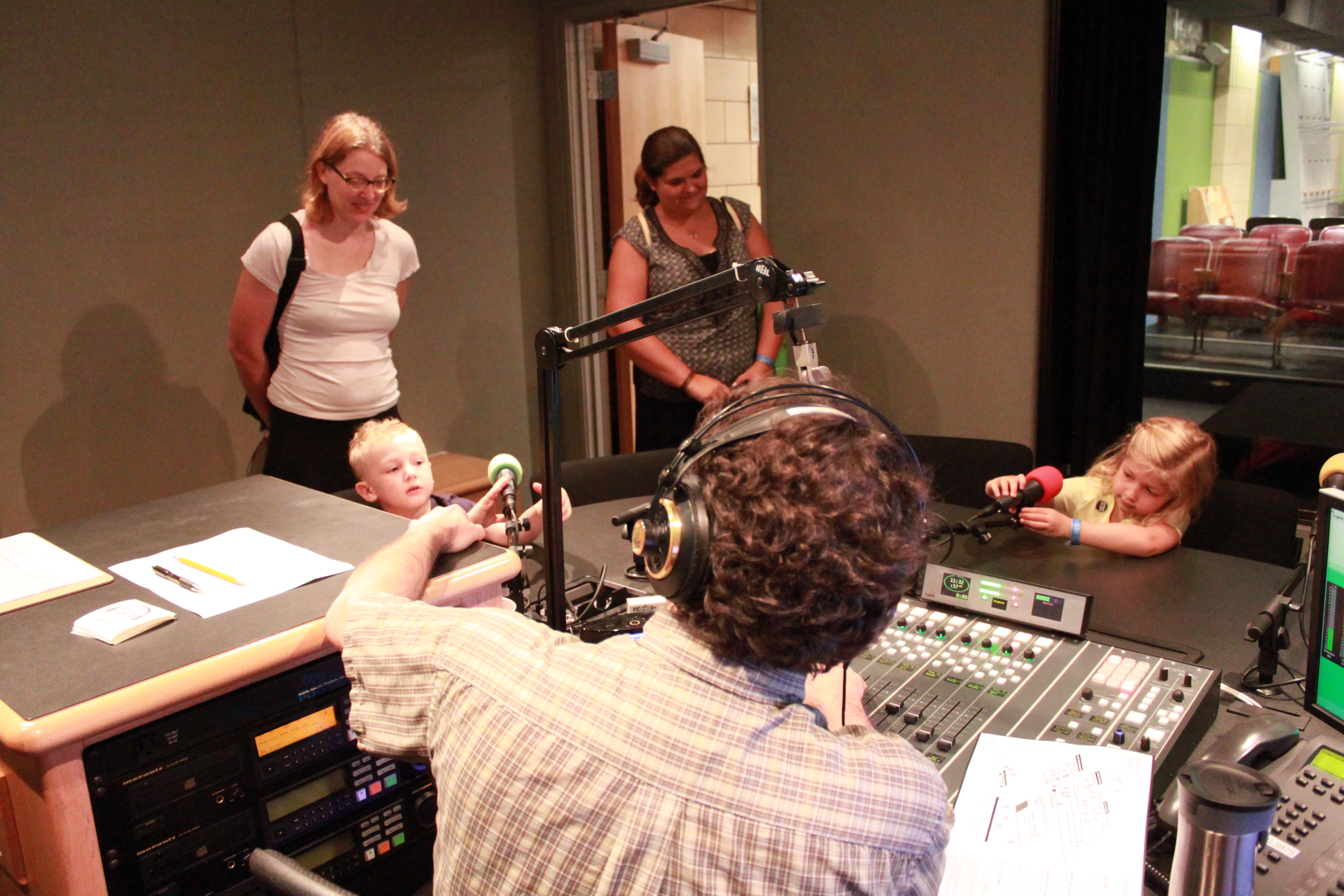 An adult wearing headphones sitting behind a microphone and a mixing board interviewing two children interacting with microphones while two other adults supervise