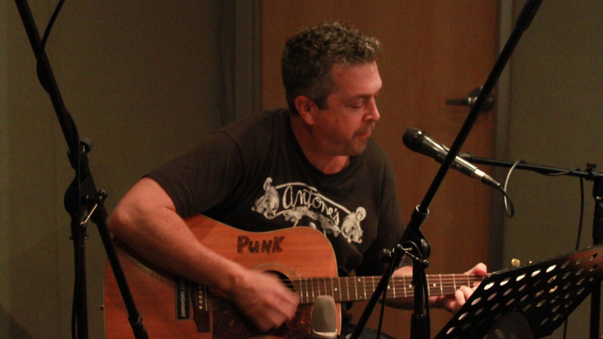 An adult playing guitar in front of a microphone and looking at sheet music on a music stand in a radio studio