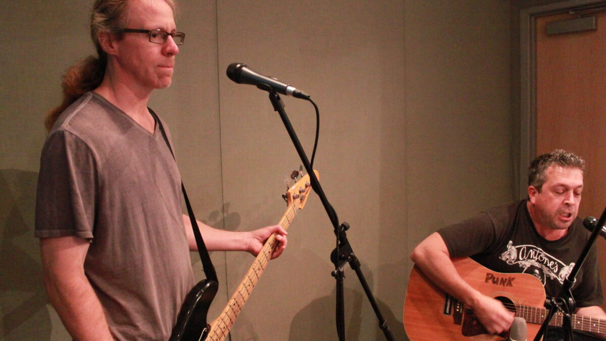 An adult playing bass in front of a microphone and an adult playing guitar while singing into a microphone in a radio studio