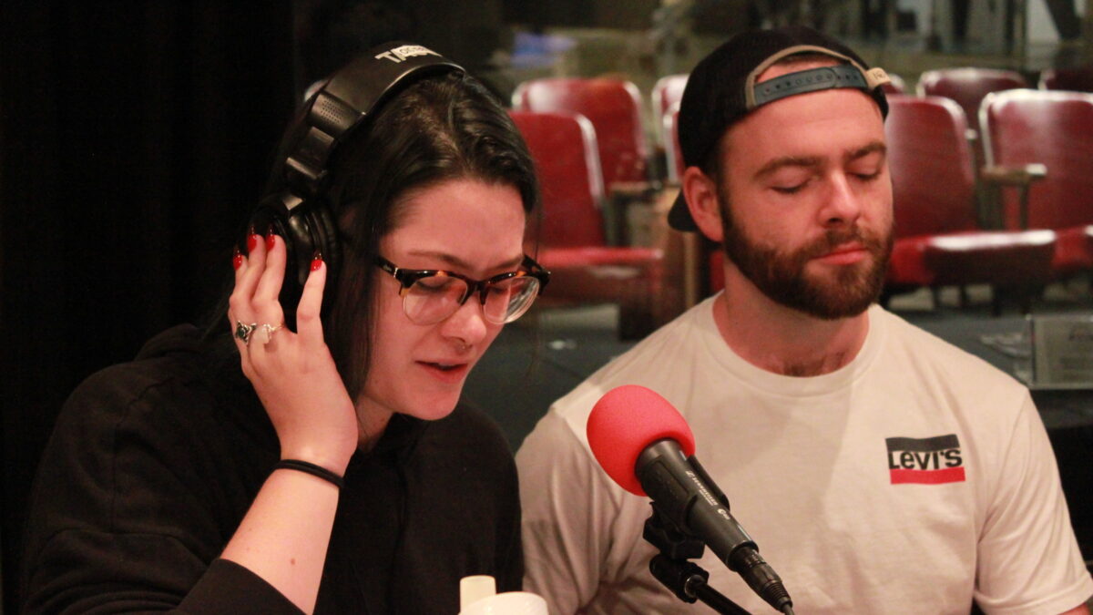 An adult wearing headphones and speaking into a microphone while holding a water bottle, while another adult listens on the side, both in a radio studio