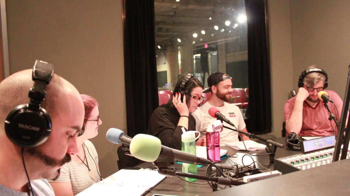 Five young adults wearing headphones sitting in front of microphones and looking down at binders and pieces of paper in a radio studio