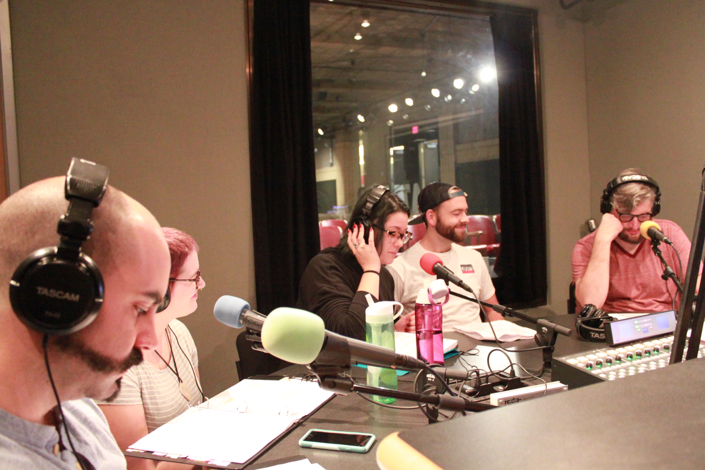Five young adults wearing headphones sitting in front of microphones and looking down at binders and pieces of paper in a radio studio