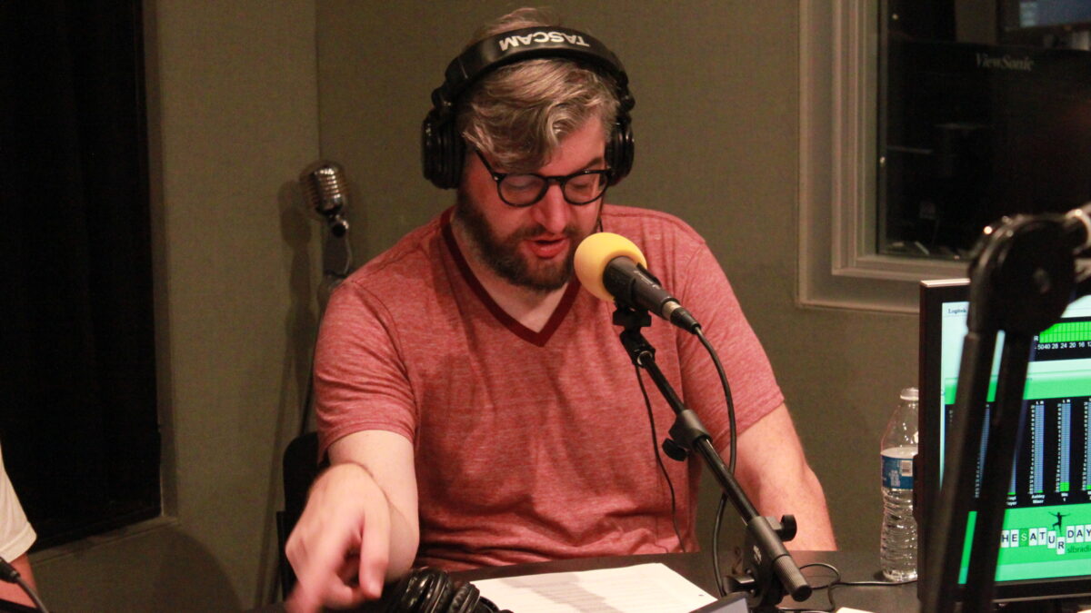 Adult wearing headphones and speaking into a microphone while looking down at a piece of paper and pointing to the side in a radio studio