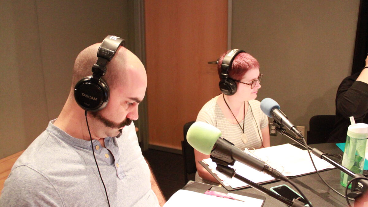 Two adults wearing headphones sitting in front of microphones and looking down at binders in a radio studio