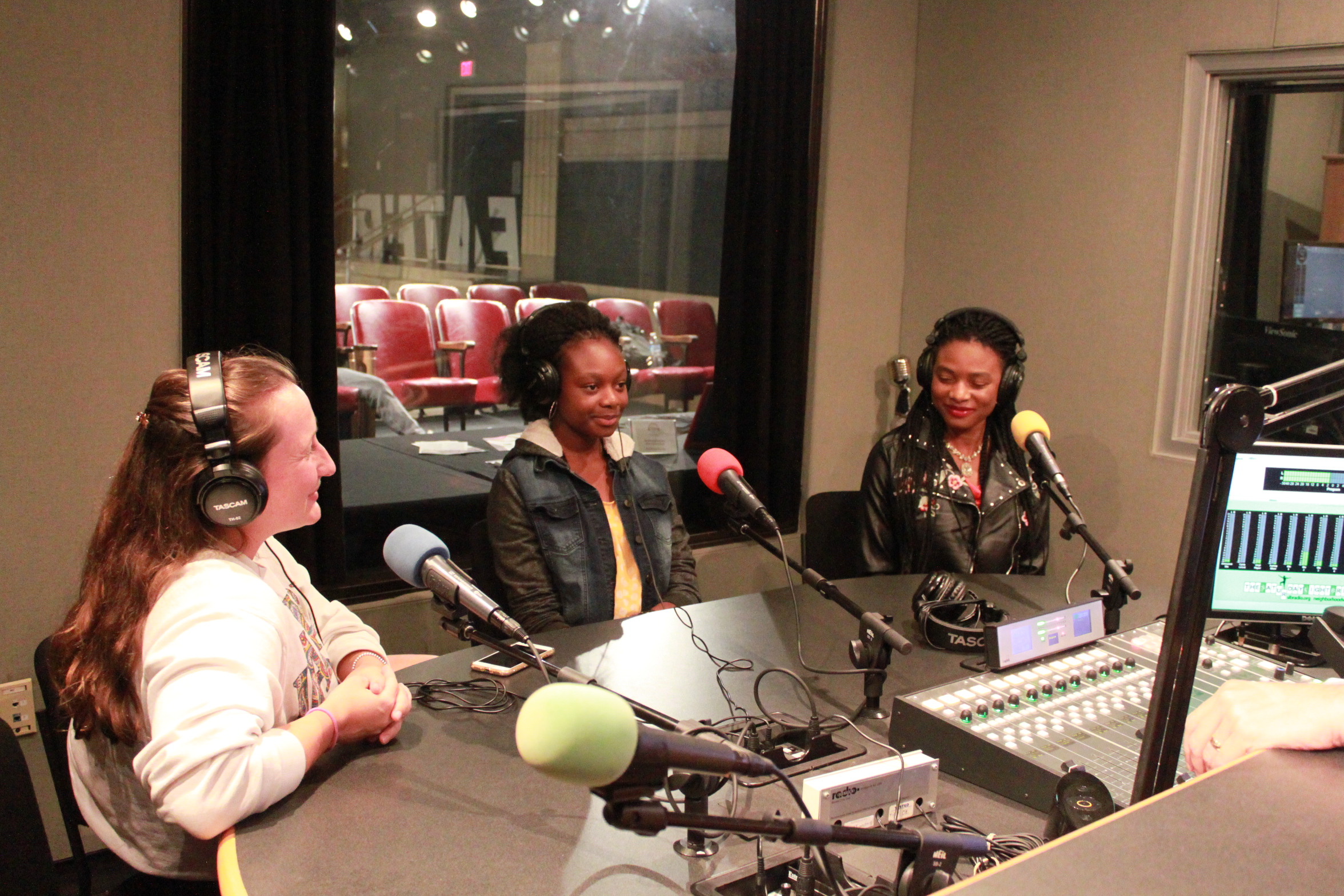 Two teens and one adult, all wearing headphones and sitting in front of microphones in a radio studio