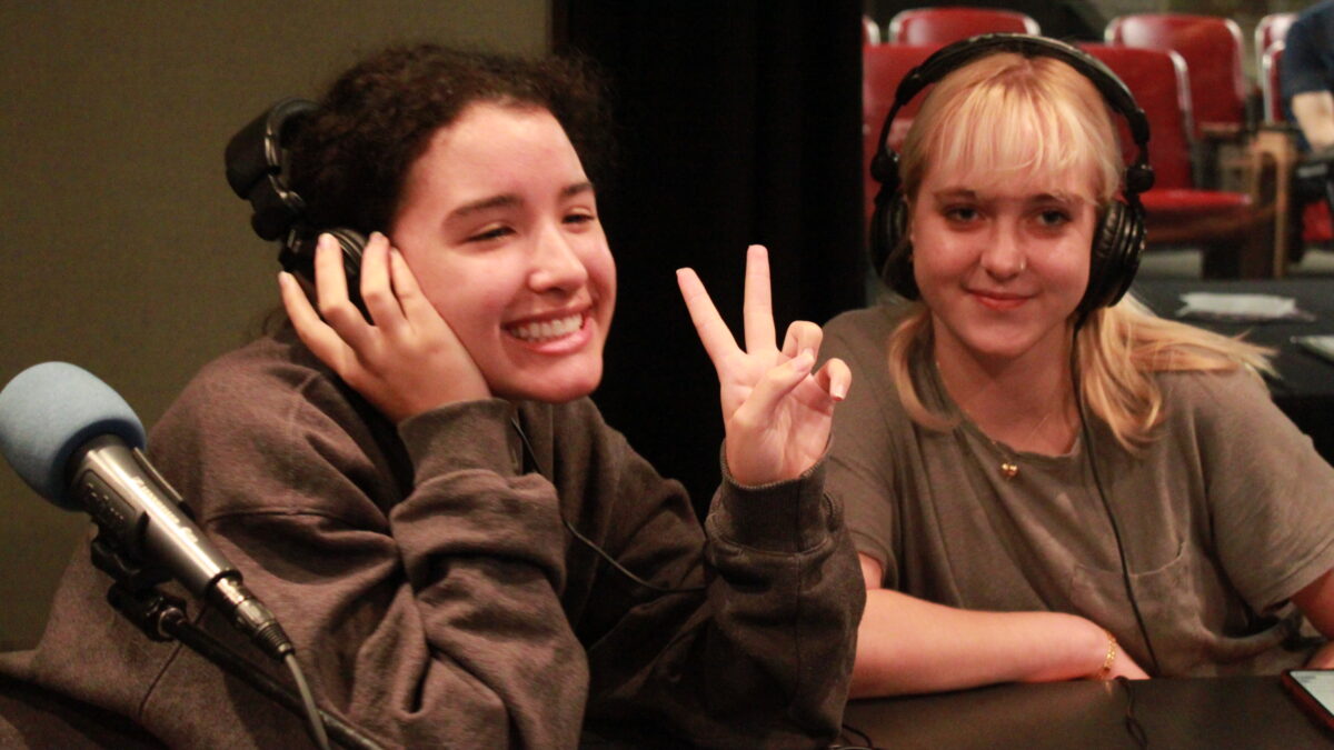 Two teen wearing headphones smiling and posing next to microphones in a radio studio