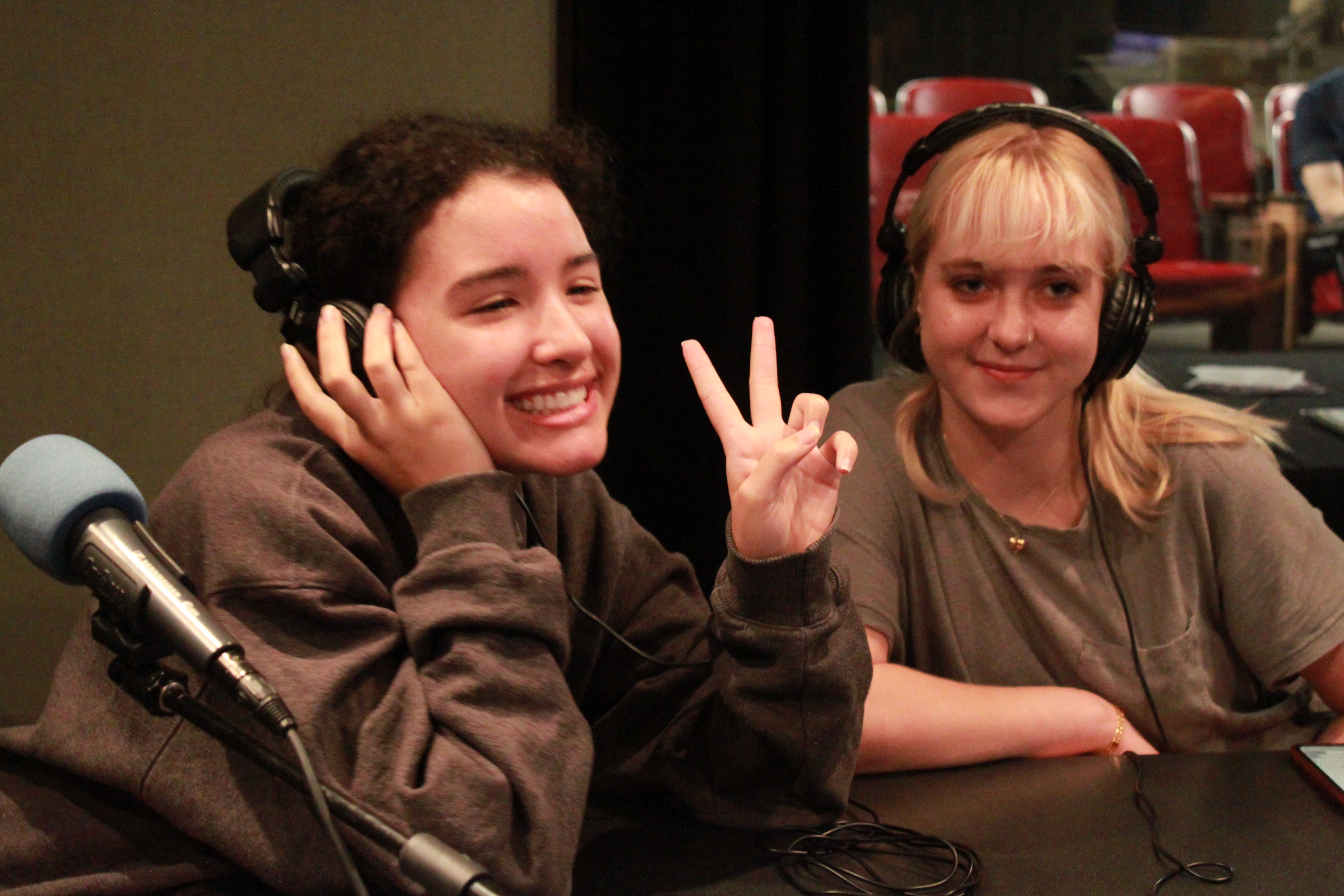 Two teen wearing headphones smiling and posing next to microphones in a radio studio