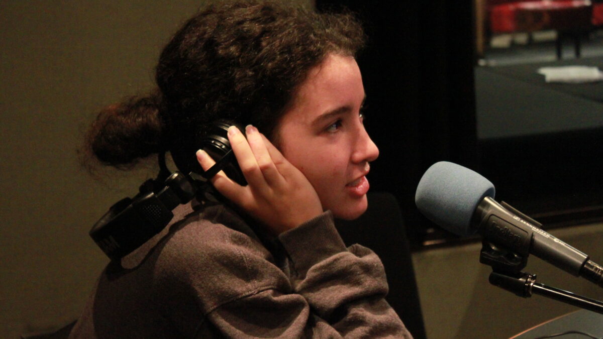Teen holding headphones up to their head and speaking into a microphone in a radio studio