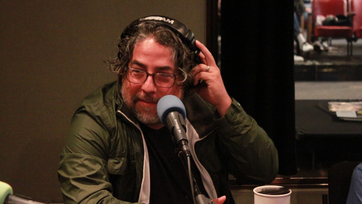 Adult wearing glasses and headphones speaking into a microphone and grasping the headphones with their hand in a radio studio