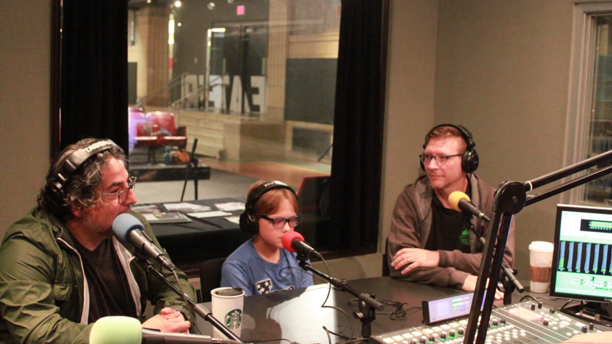 Two adults and one youth, all wearing glasses and headphones and speaking into microphones in a radio studio