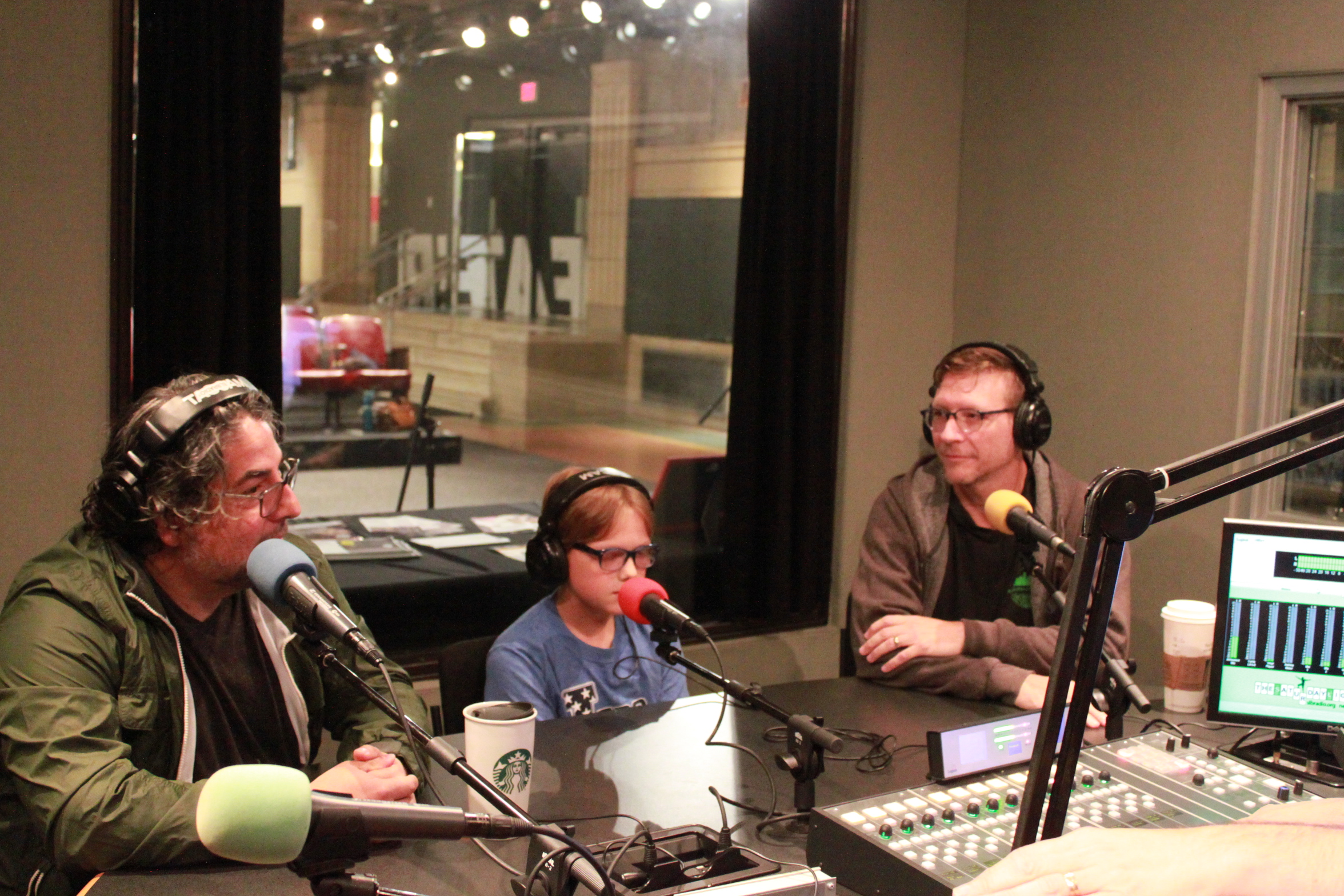 Two adults and one youth, all wearing glasses and headphones and speaking into microphones in a radio studio