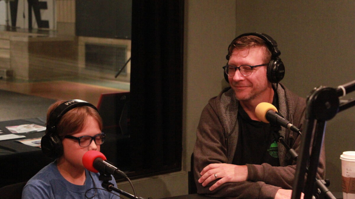 One adult and one youth, both wearing glasses and headphones and speaking into microphones in a radio studio