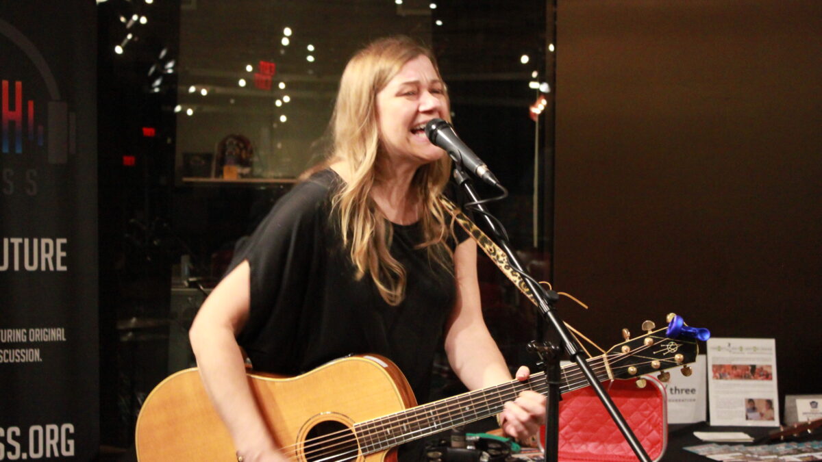 Adult singing into a microphone and playing guitar outside a radio studio