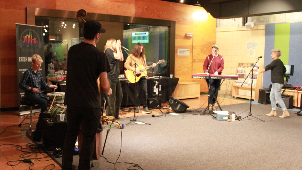 Six adult musicians performing outside a radio studio, one playing upright bass, one playing drums, one playing guitar, one playing keyboard, and three singing