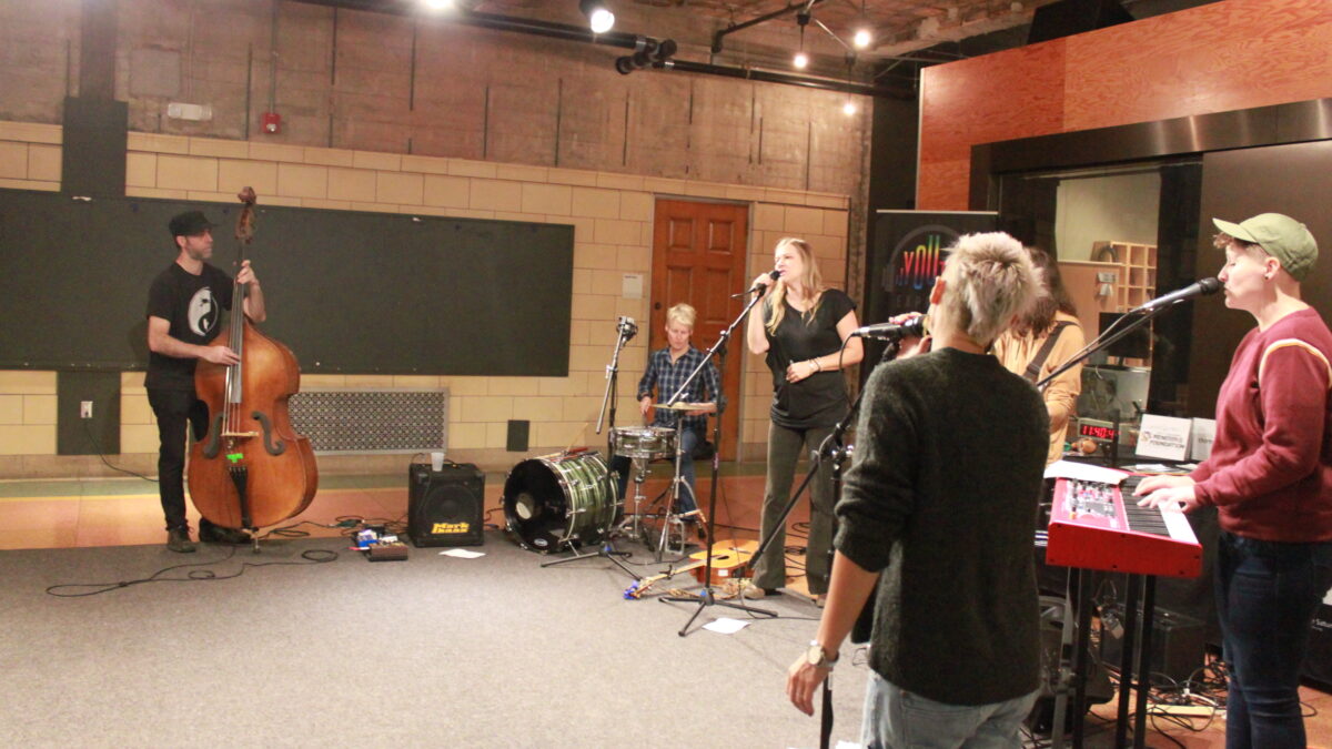 Six adult musicians performing outside a radio studio, one playing upright bass, one playing drums, one playing guitar, one playing keyboard, and three singing
