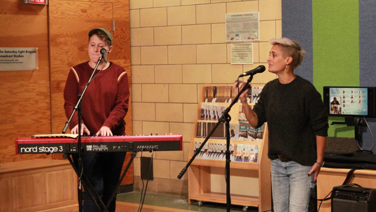 An adult playing piano keyboard and an adult singing while playing a shaker outside a radio studio