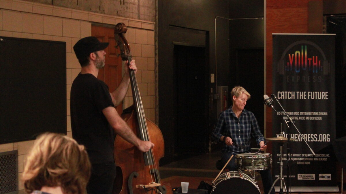 An adult playing upright bass and an adult playing drums outside a radio studio