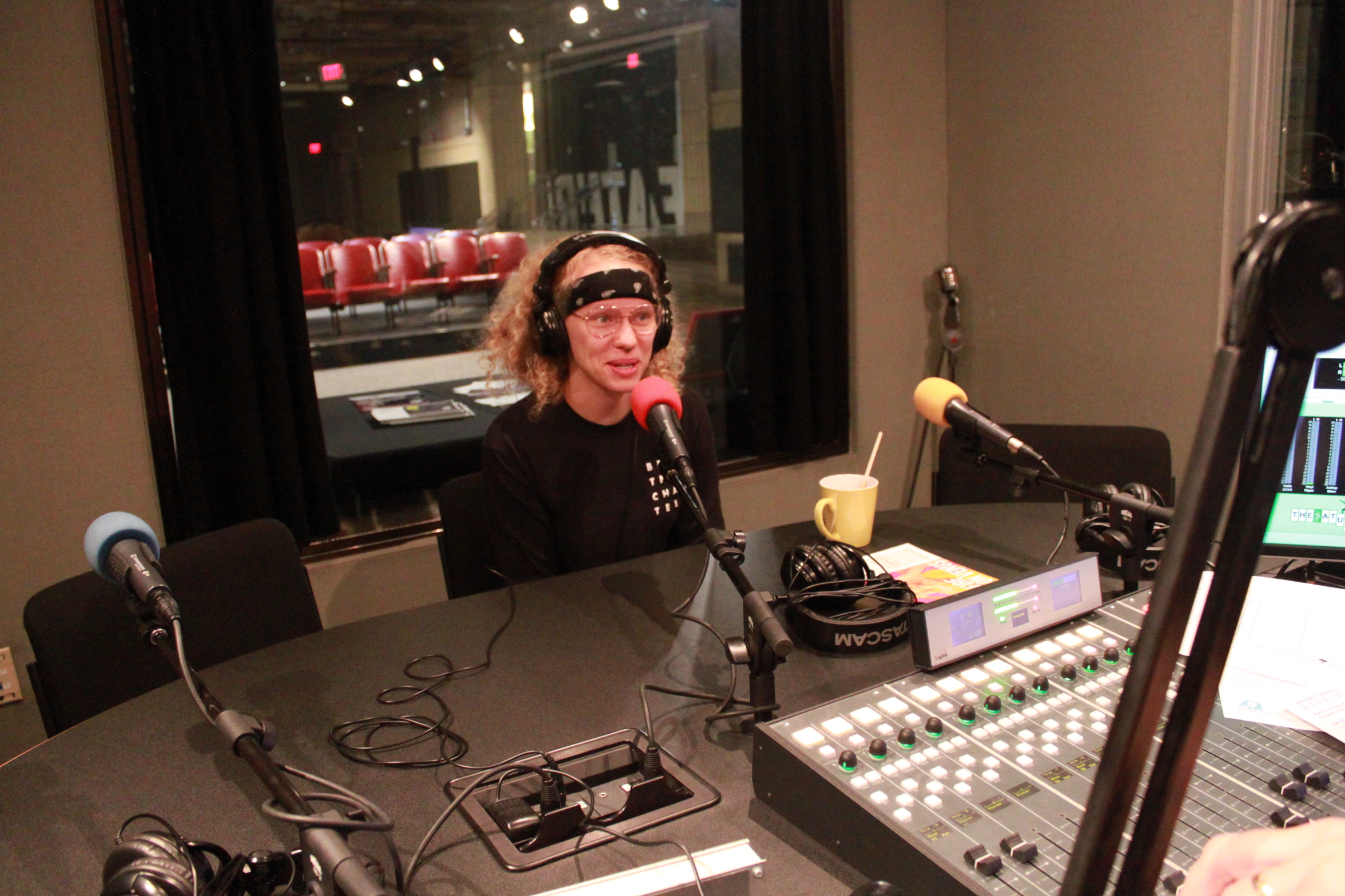 Adult wearing glasses, a head scarf, and headphones speaking into a microphone in a radio studio