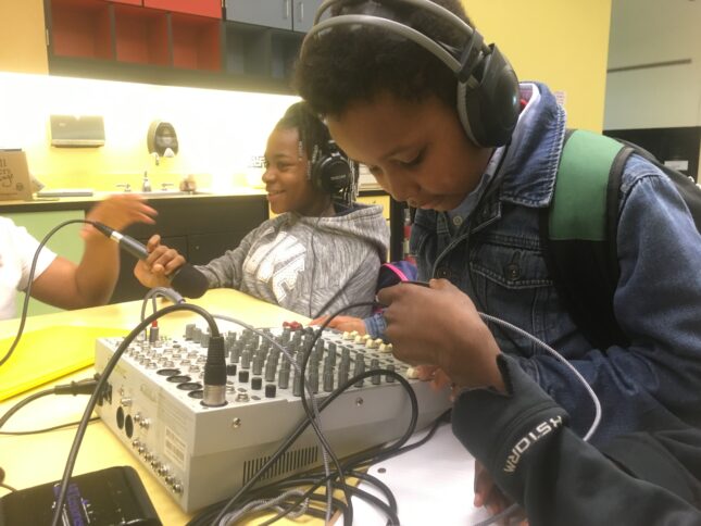 A youth wearing headphones interacting with a mixing board and a youth wearing headphones holding a microphone upside down