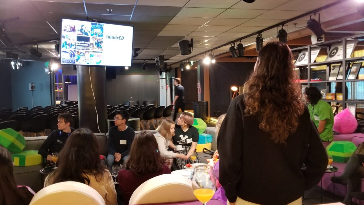 A group of young adults sitting on purple and gray couches looking at a tv screen