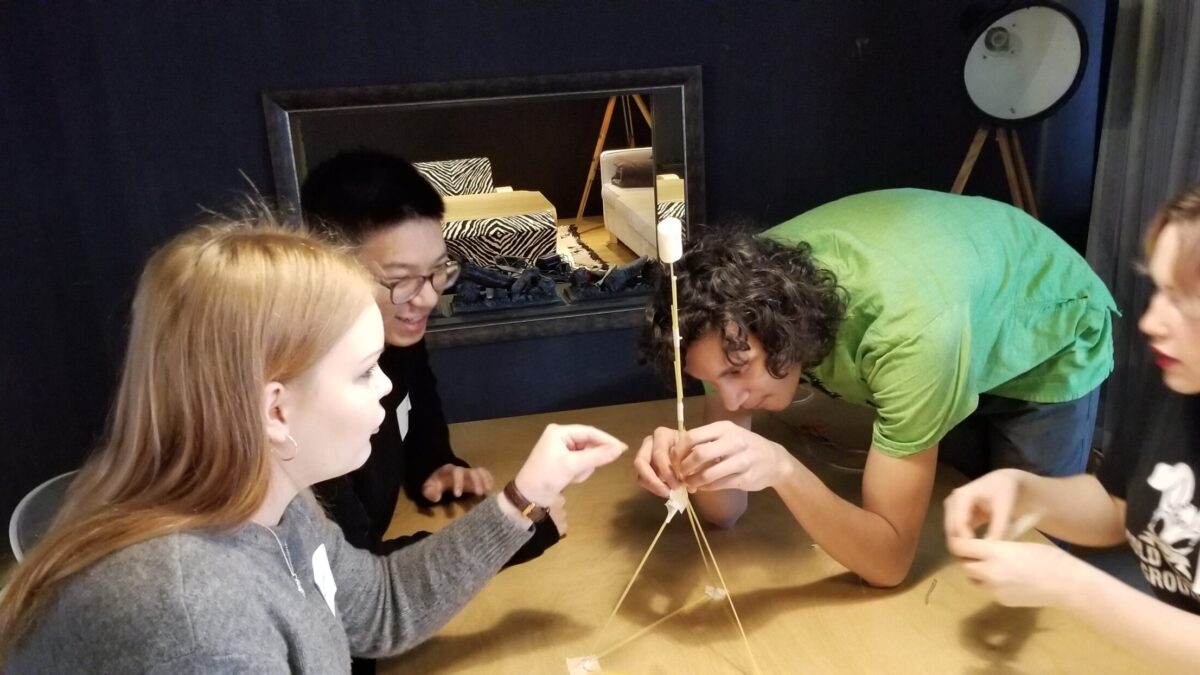Four young adults building a structure out of wooden sticks and marshmallows on a light brown table