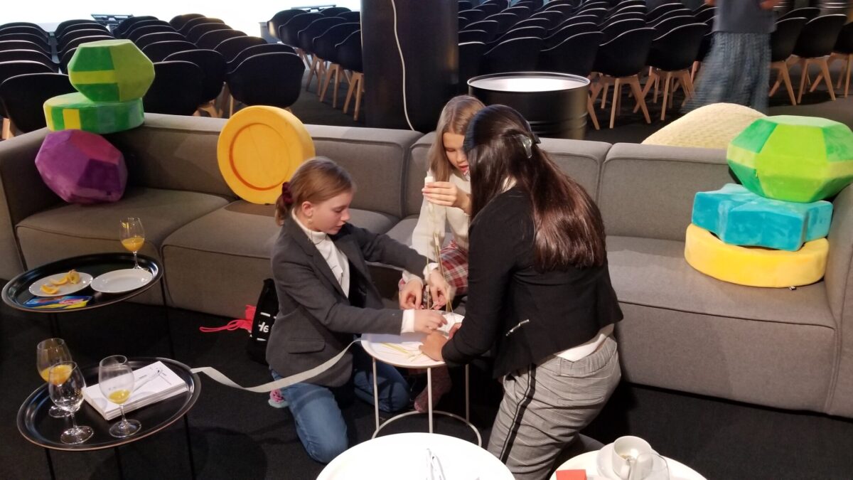 Three young adults building a sculpture out of wooden sticks and marshmallows on a small white table in front of a long gray couch