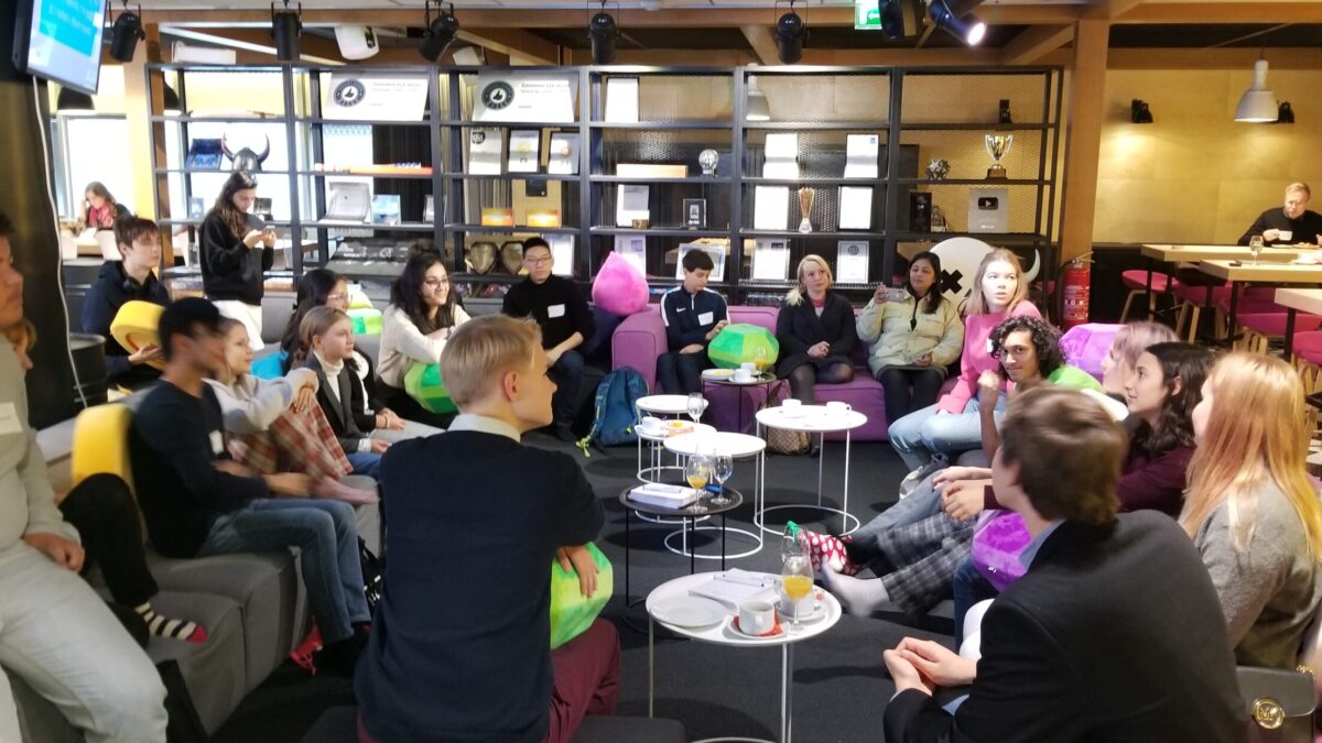 A large group of young adults sitting on gray and purple couches around small white tables having a discussion