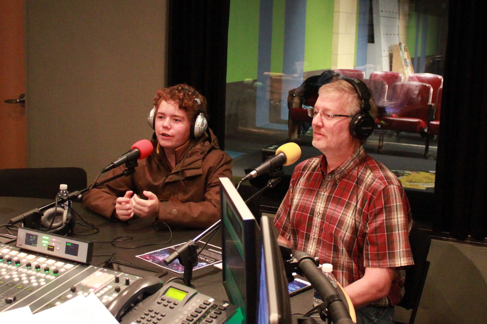 An adult and a youth both wearing headphones and speaking into microphones in a radio studio